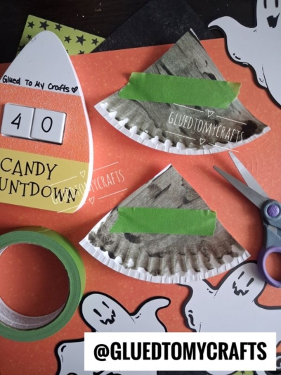 Halloween-themed craft scene with paper ghosts, orange cardstock, a painted paper plate cut in half, green tape, scissors, and a large candy corn countdown sign—perfect inspiration for Halloween Crafts for Kids. @GLUEDTOMYCRAFTS is labeled at the bottom.