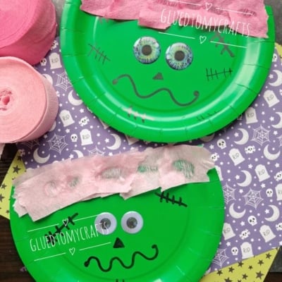Two green paper plates decorated as Frankenstein heads with pink crepe paper hair, googly eyes, and black marker faces, placed on top of purple Halloween-themed paper. Pink crepe paper rolls are on the side.