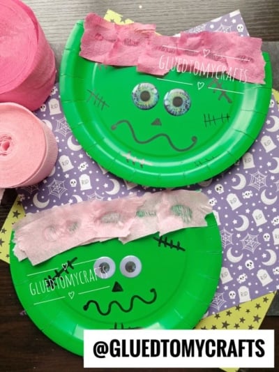 Two green paper plates decorated as Frankenstein heads with pink crepe paper hair, googly eyes, and black marker faces, placed on top of purple Halloween-themed paper. Pink crepe paper rolls are on the side.