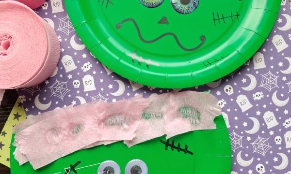 Two green paper plates decorated as Frankenstein heads with pink crepe paper hair, googly eyes, and black marker faces, placed on top of purple Halloween-themed paper. Pink crepe paper rolls are on the side.