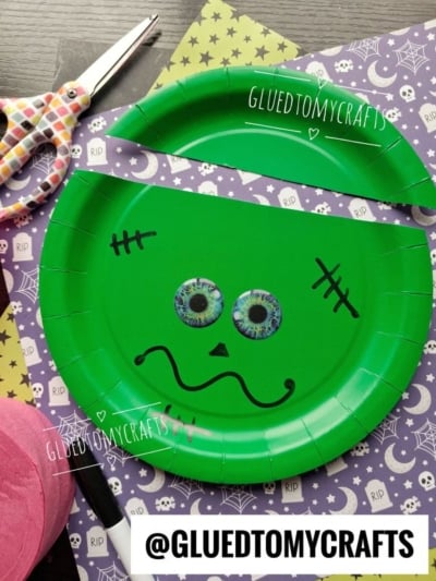 A green paper plate decorated as Frankensteins face with googly eyes, black marker features, and background Halloween-themed paper and scissors.