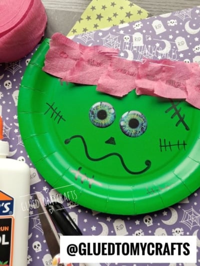 A green paper plate craft resembling Frankenstein with googly eyes and pink crepe hair sits on a purple Halloween-themed paper, surrounded by glue, scissors, and crafting supplies. @GLUEDTOMYCRAFTS is at the bottom.