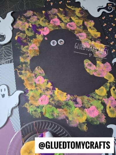 A black paper features a ghost shape surrounded by colorful, sponged paint. Googly eyes are attached to the ghost. Ghost decorations and art supplies are around the paper. Text reads @GLUEDTOMYCRAFTS.