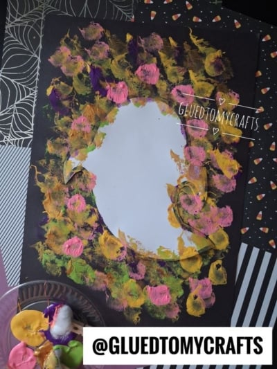 A black paper with a white oval center, surrounded by dabs of yellow, pink, and purple paint, sits on patterned paper. Nearby are paint colors and the text @GLUEDTOMYCRAFTS..
