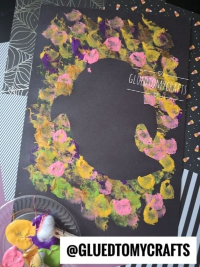 A black paper with a penguin silhouette outlined in dabs of yellow, green, pink, and purple paint. Paint and cotton swabs are on a plate nearby. @GLUEDTOMYCRAFTS text appears at the bottom.