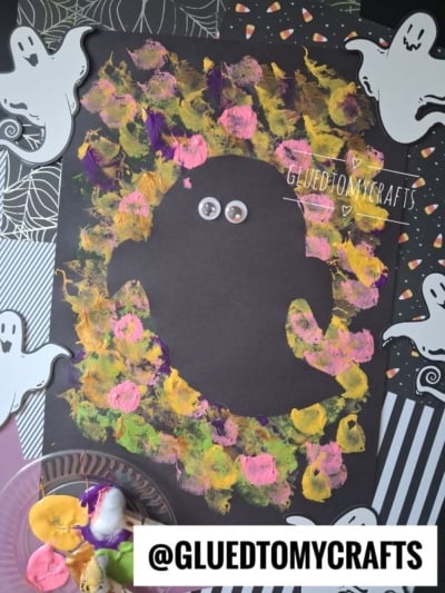 A black paper ghost surrounded by colorful sponge-painted spots, with googly eyes and paper cut-out ghosts in the background. Paint palette with brushes is seen in the corner.