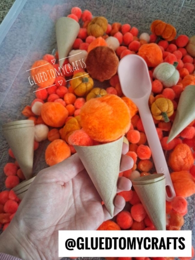 A hand holds a cardboard cone filled with orange pom-poms, inviting imaginative pom pom ice cream play above a bin of orange and brown pom-poms, mini cones, and a pink plastic scoop. Text: @GLUEDTOMYCRAFTS.