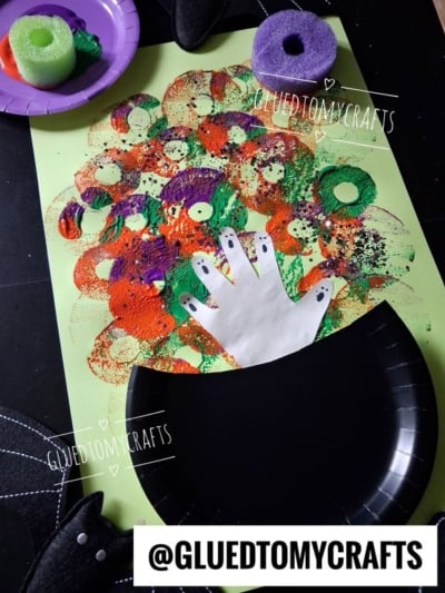 A Halloween craft featuring a paper cauldron, a handprint as a hand coming out, and colorful paint circles as bubbles on green paper. Pool noodles and craft supplies are around the artwork.