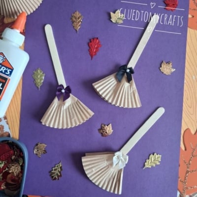 Three popsicle stick dolls with cupcake liner dresses and bows are arranged on purple paper, surrounded by autumn leaf decorations, Elmers glue, and a container of embellishments. @GLUEDTOMYCRAFTS is written at the bottom.