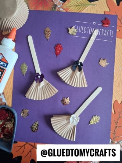 Three popsicle stick dolls with cupcake liner dresses and bows are arranged on purple paper, surrounded by autumn leaf decorations, Elmers glue, and a container of embellishments. @GLUEDTOMYCRAFTS is written at the bottom.
