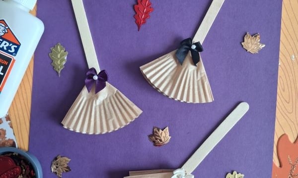 Three popsicle stick dolls with cupcake liner dresses and bows are arranged on purple paper, surrounded by autumn leaf decorations, Elmers glue, and a container of embellishments. @GLUEDTOMYCRAFTS is written at the bottom.