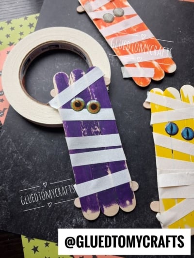 Three craft stick mummies are decorated with colored stripes, googly eyes, and white tape, displayed on a black surface with a roll of tape and colored paper. @GLUEDTOMYCRAFTS watermark appears twice.