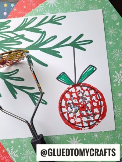 A painted Christmas ornament craft made with a potato masher stamped craft technique, using red paint and glitter on white paper, with pine branch drawings and a green patterned background.