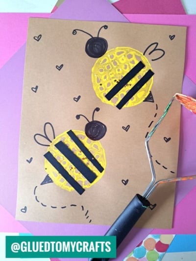 A brown paper shows two bumblebees painted with yellow and black, accented with hearts and a potato masher tool, on top of colorful paper sheets—perfect for a fun potato masher stamped craft. Text reads @GLUEDTOMYCRAFTS.