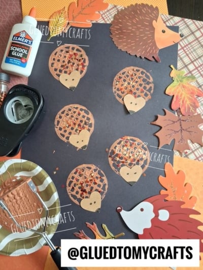 A black paper displays hedgehog crafts made from brown circles and leaf cutouts, surrounded by glue, a hole punch, a paper plate with markers, and autumn-themed decorations. A @GLUEDTOMYCRAFTS tag is visible.