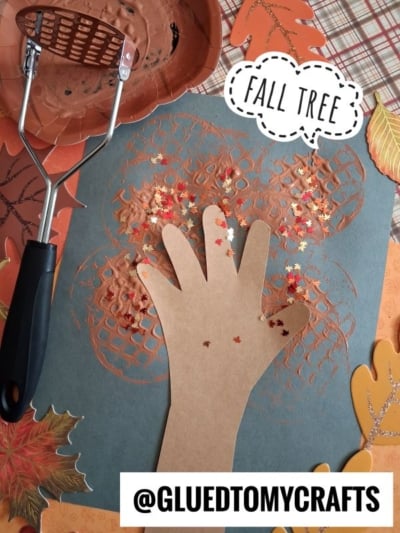 A fall tree craft with a paper hand as the tree trunk, orange paint patterns, scattered leaf-shaped confetti, and a potato masher. FALL TREE and @GLUEDTOMYCRAFTS text included. Autumn leaves surround the artwork.