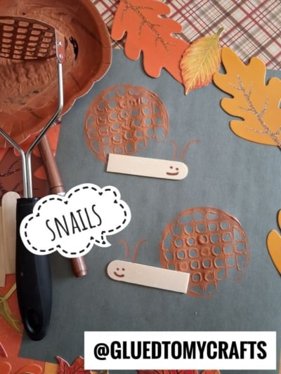 A fall-themed snail craft using mashed potato mashers to create snail shells with brown paint, popsicle sticks as snail bodies, and paper leaves as decoration. The word SNAILS and @GLUEDTOMYCRAFTS appear.