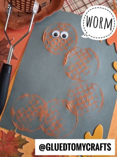 A craft project with orange paint circles forming a worm shape on green paper, googly eyes, labeled WORM. A potato masher with paint and autumn decorations are nearby. Text: @GLUEDTOMYCRAFTS..