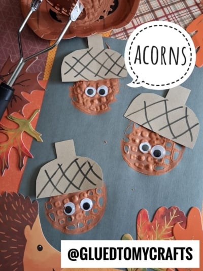 Three paper acorn crafts with googly eyes are displayed on a table with fall-themed decorations. The word ACORNS appears in a speech bubble. The Instagram handle @GLUEDTOMYCRAFTS is shown at the bottom.