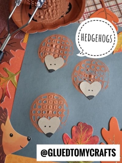 Three brown waffle-like paper hedgehogs with heart-shaped faces are on gray paper, surrounded by autumn leaves and craft tools. A speech bubble says HEDGEHOGS. The handle @GLUEDTOMYCRAFTS is at the bottom.