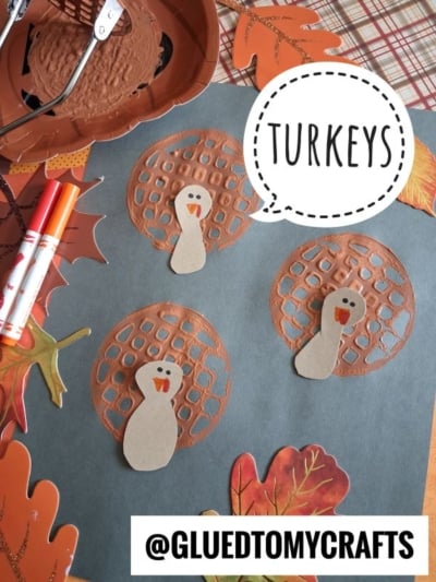 Three paper turkeys made with brown painted circles and tan bodies are glued to dark paper, surrounded by colorful paper leaves and markers. The word TURKEYS is in a speech bubble.