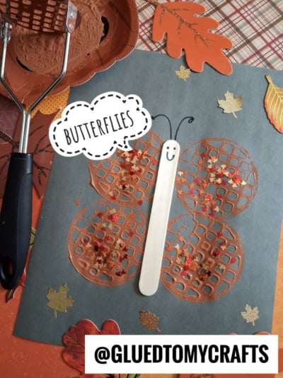 A fall-themed butterfly craft made with brown painted potato masher prints, a craft stick body, smiley face, and red-gold glitter, on a blue paper with leaf decorations. @GLUEDTOMYCRAFTS at the bottom.
