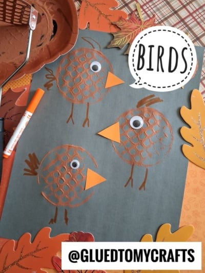 Three brown, honeycomb-textured bird crafts with googly eyes and orange beaks on black paper. Fall leaves and markers are scattered around. BIRDS is written in a speech bubble.