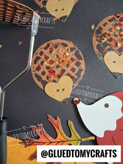 Hedgehog crafts made from brown paint, paper hearts for faces, and autumn leaf confetti on black paper. A potato masher and paper leaves are nearby. @gluedtomycrafts watermark appears on the image.