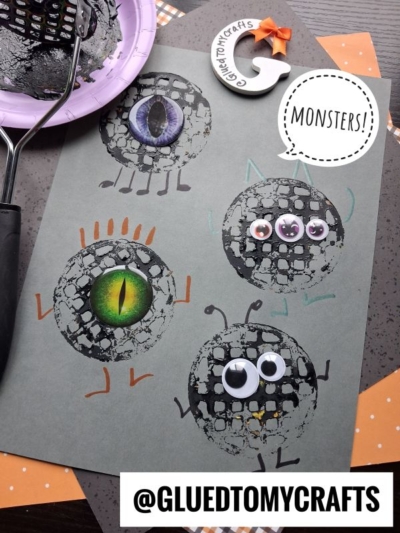 Four black, round monster faces with different googly eyes and colorful details are painted on gray paper—perfect for inspiring fun Halloween potato masher crafts. Nearby are a paint roller, a crescent moon clay craft, and a tag reading @gluedtomycrafts.