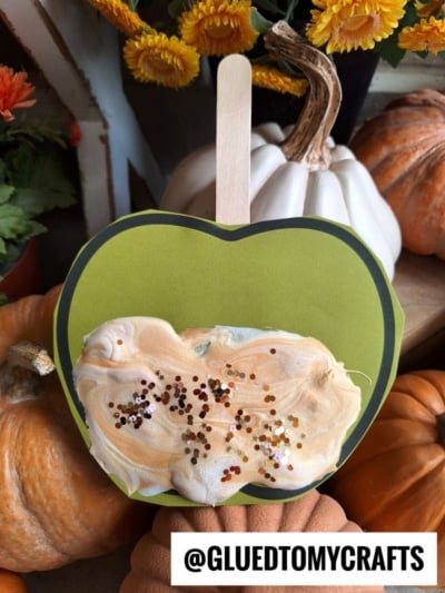 A green paper apple on a stick with swirled puffy paint “caramel” frosting and sprinkles, surrounded by pumpkins and autumn flowers. Text at bottom: @GLUEDTOMYCRAFTS.