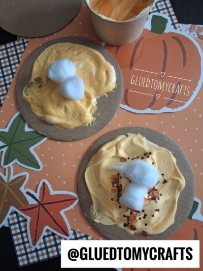 Two round cardboard pieces topped with orange cream, cotton balls, and sprinkles sit on autumn-themed paper with leaves and a pumpkin. Perfect for FALL CRAFTS FOR KIDS, this cheerful setup also features a cup of orange cream and the handle @GLUEDTOMYCRAFTS.