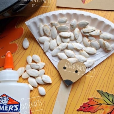 A paper plate hedgehog craft with pumpkin seeds, brown paper, glue, scissors, and fall leaves on a wood background. The image has GLUEDTOMYCRAFTS and @GLUEDTOMYCRAFTS watermarks.