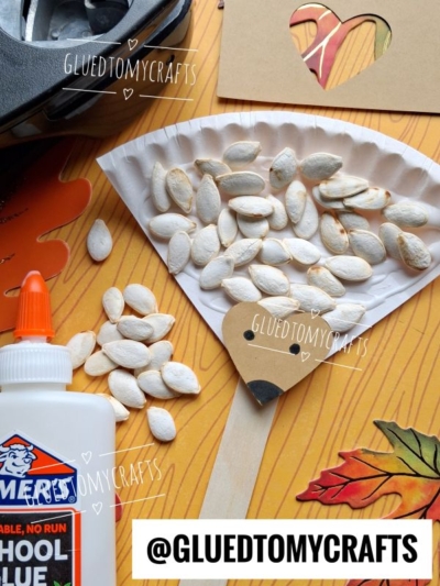 A paper plate broom craft with pumpkin seeds, brown paper, glue, scissors, and fall leaves on a wood background. The image has GLUEDTOMYCRAFTS and @GLUEDTOMYCRAFTS watermarks.