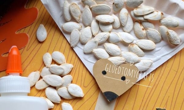 A paper plate hedgehog craft with pumpkin seeds, brown paper, glue, scissors, and fall leaves on a wood background. The image has GLUEDTOMYCRAFTS and @GLUEDTOMYCRAFTS watermarks.