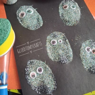 Five glittery, oval shapes with googly eyes are glued to black paper, creating a fun monster craft. Craft supplies and the handle of a sponge brush are visible nearby. @GLUETOMYCRAFTS is displayed at the bottom.