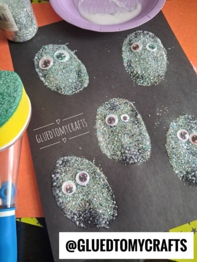 Five glittery, oval shapes with googly eyes are glued to black paper, creating a fun monster craft. Craft supplies and the handle of a sponge brush are visible nearby. @GLUETOMYCRAFTS is displayed at the bottom.