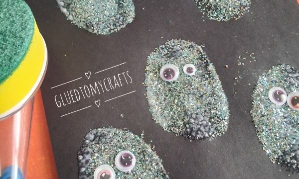 Five glittery, oval shapes with googly eyes are glued to black paper, creating a fun monster craft. Craft supplies and the handle of a sponge brush are visible nearby. @GLUETOMYCRAFTS is displayed at the bottom.
