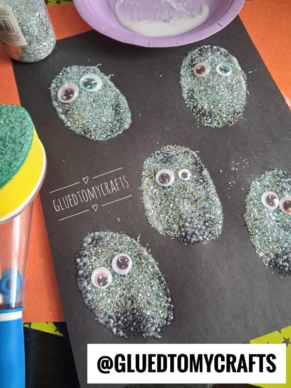Five glittery, oval shapes with googly eyes are glued to black paper, creating a fun monster craft. Craft supplies and the handle of a sponge brush are visible nearby. @GLUETOMYCRAFTS is displayed at the bottom.