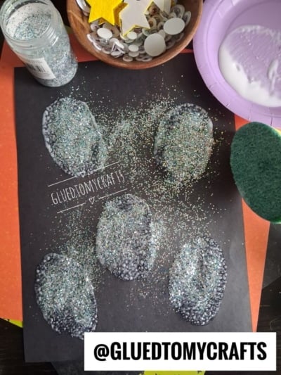 A black paper with five gray ovals covered in glitter, surrounded by craft supplies like foam shapes, a jar of glitter, a bowl of glue, and a green sponge. @GLUEDTOMYCRAFTS is displayed at the bottom.