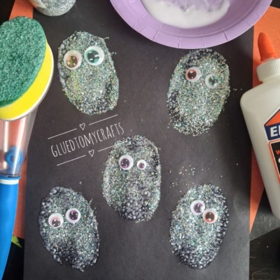 Five oval shapes covered in glitter and googly eyes are glued to black paper. Craft supplies like glue, a sponge, and a bowl of liquid can be seen nearby. @GLUEDTOMYCRAFTS is printed on the image.