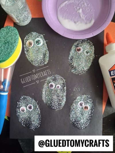 Five oval shapes covered in glitter and googly eyes are glued to black paper. Craft supplies like glue, a sponge, and a bowl of liquid can be seen nearby. @GLUEDTOMYCRAFTS is printed on the image.
