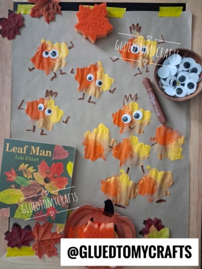 Crafted leaf shapes painted in autumn colors with googly eyes are arranged on a table. Showcasing one of the best fall crafts for kids, the scene also features extra googly eyes, a glue stick, the book Leaf Man, and festive fall-themed decorations.
