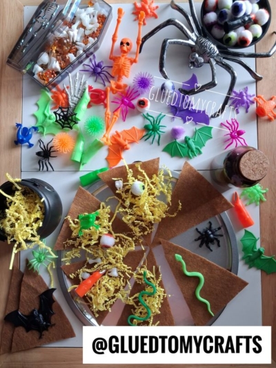 Colorful Halloween-themed crafts and toys, including plastic spiders, bats, skeletons, and snakes, are arranged on a table with brown paper triangles, yellow shredded paper, and candies—perfect for inspiring FALL THEMED SENSORY BIN IDEAS. @GLUETOMYCRAFTS text is visible.