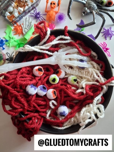 A bowl of red and white craft yarn “spaghetti” with fake eyeballs and a skeleton hand, surrounded by playful plastic Halloween decorations. Text: @GLUEDTOMYCRAFTS.