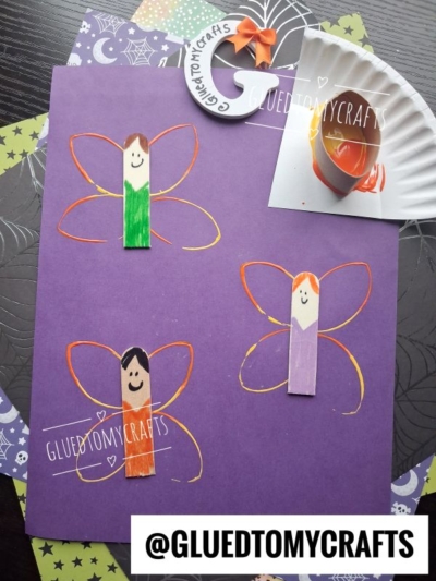 Three fairy crafts made from clothespins and paper are arranged on purple paper, perfect examples of FALL CRAFTS FOR KIDS. Each fairy has colorful wings and a smiling face, bringing cheerful autumn vibes. A partial orange and white craft is in the corner. The handle @GLUEDTOMYCRAFTS is visible.
