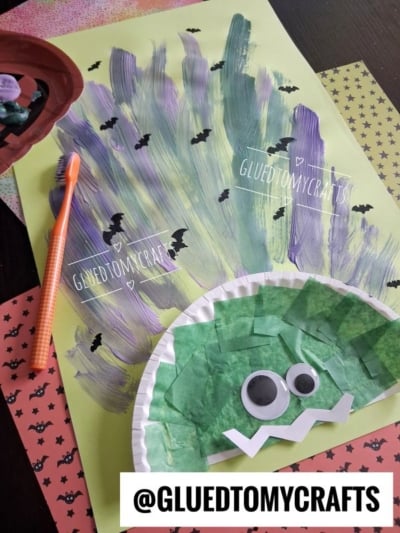 A Halloween craft featuring a Frankenstein face made of green paper and googly eyes on a plate, with painted purple and green strokes and bat stickers in the background. An orange toothbrush and craft materials are nearby.