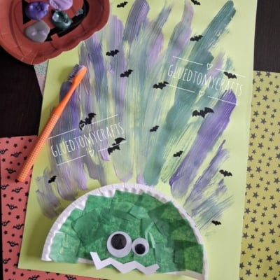 A kids craft project of a troll with purple and green painted hair, googly eyes, and a tissue paper face on green paper. Halloween-themed papers and a bowl of erasers are nearby. @GLUEDTOMYCRAFTS watermark is present.