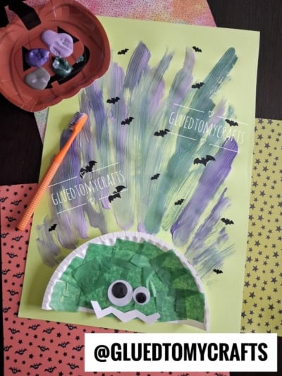 A kids craft project of a troll with purple and green painted hair, googly eyes, and a tissue paper face on green paper. Halloween-themed papers and a bowl of erasers are nearby. @GLUEDTOMYCRAFTS watermark is present.