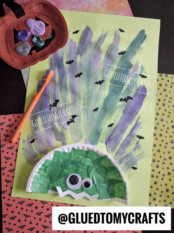A kids craft project of a troll with purple and green painted hair, googly eyes, and a tissue paper face on green paper. Halloween-themed papers and a bowl of erasers are nearby. @GLUEDTOMYCRAFTS watermark is present.