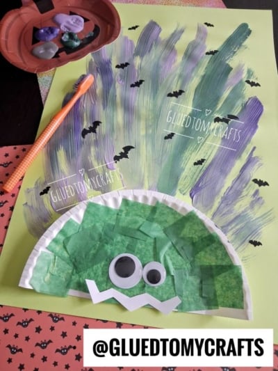 A Frankenstein craft made from a paper plate with green tissue, googly eyes, and zigzag mouth sits on painted paper with bats, beside crystals, a pencil, and a @gluedtomycrafts label.